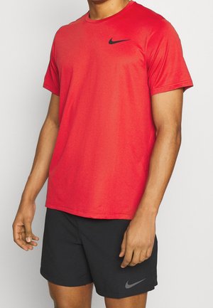 Uomo con maglietta rossa a maniche corte Nike e pantaloncini sportivi neri Nike, in piedi davanti a uno sfondo chiaro semplice.