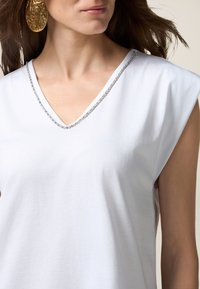 T-shirt in cotone bianco con scollatura a V, caratterizzata da un bordo decorato con strass argentati. Design senza maniche con una texture liscia.