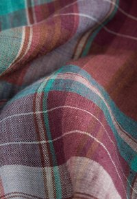 Gros plan sur un tissu texturé avec un motif à carreaux tissé en bordeaux, bleu sarcelle, blanc et marron, montrant des plis et des fils détaillés.