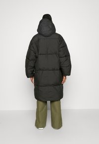 Lange zwarte pufferjas met een oversized pasvorm, capuchon en doorgestikt ontwerp. Een matte textuur, gecombineerd met olijfgroene wijde broek en witte sneakers.