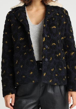 Kvinde iført en sort tekstureret uldjakke med gule pletter, en hvid fåreskindskrave, en lysegrå ribbet top og sorte læderbukser.