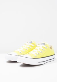 Żółte płócienne trampki z białymi gumowymi podeszwami, czarnymi paskami i srebrnymi oczkami. Posiadają białe sznurówki oraz okrągły nos.