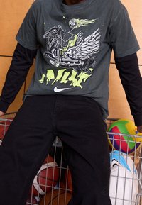 Camiseta gris gráfica con un diseño en verde y blanco que presenta un caballo alado, combinada con pantalones negros, situada frente a un fondo de varias pelotas deportivas.