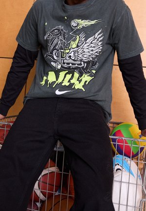 Grafisk grå t-shirt med grön och vit design som föreställer en bevingad häst, i kombination med svarta byxor, mot en bakgrund av olika sportbollar.