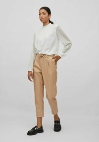 Bela bluza z dolgimi rokavi z teksturiranimi chevron vzorci, kombinirana z bež hlačami v usklajenem kroju in črnimi loaferji z zlatimi dodatki.