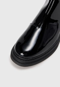 Sorte patentlær ankle boot med rund tå, en glat finish og en tyk sort gummisål. Syningsdetalje foran.