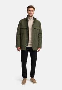 Olijfgroene jas met twee borstzakken, beige gebreide trui, zwarte broek en tan schoenen. Model staat tegen een effen achtergrond.