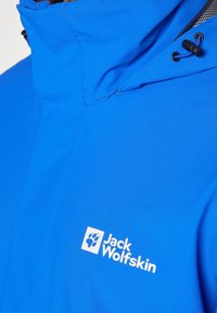 Veste extérieure bleue avec capuche réglable équipée d'un cordon à bascule et logo blanc Jack Wolfskin sur la poitrine.