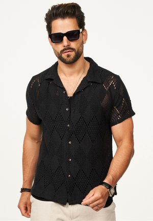Black short-sleeve shirt made of open-knit fabric with diamond patterns, featuring a collar and brown buttons. Model wears sunglasses.