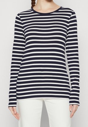 Vrouw draagt een marineblauw-wit horizontaal gestreept shirt met lange mouwen en een witte broek, staand tegen een effen achtergrond.