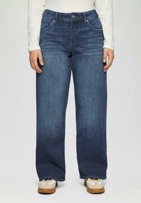 Donkerblauwe denim wide-leg jeans met een middelhoge taille, vijf zakken en contrasterende stiksels. Gecombineerd met tan en witte sneakers.