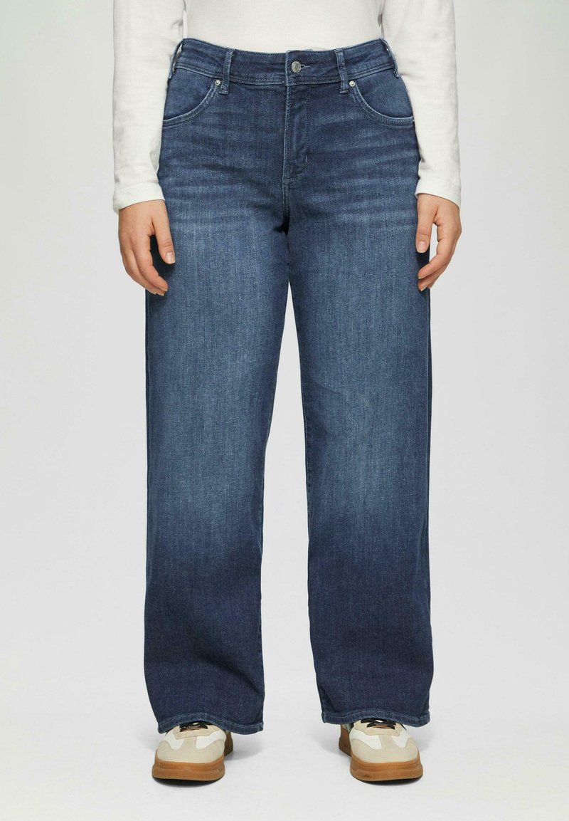 Dunkelblaue Denim-Jeans mit weit geschnittenem Bein, mittlerer Leibhöhe, fünf Taschen und kontrastierenden Nähten. Kombiniert mit beige-weißen Turnschuhen.