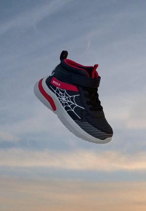 Une basket montante noire et rouge ornée d’un motif de toile d’araignée blanc flotte sur fond de ciel nuageux.