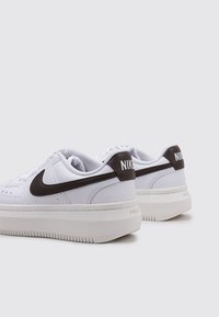 Zapatillas Nike con parte superior de cuero blanco, logo en forma de swoosh de color marrón oscuro, diseño de perfil bajo y suela de goma texturizada.
