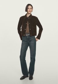 Kvinde med kort hår iført en mørkebrun fløjlskajak, brun skjorte, sort bælte med nitter, mørk jeans og sorte loafers, som står mod en ensfarvet baggrund.