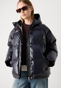 Námořnická puffer bunda s lesklým povrchem, kapucí a zapínáním na zip. Má tlačítka na stisknutí a nastavitelnou šňůrku. Nositá přes hnědý top.