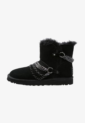 Bottine en daim noir avec doublure duveteuse et chaînes argentées décoratives entourant la tige et la zone du pied.