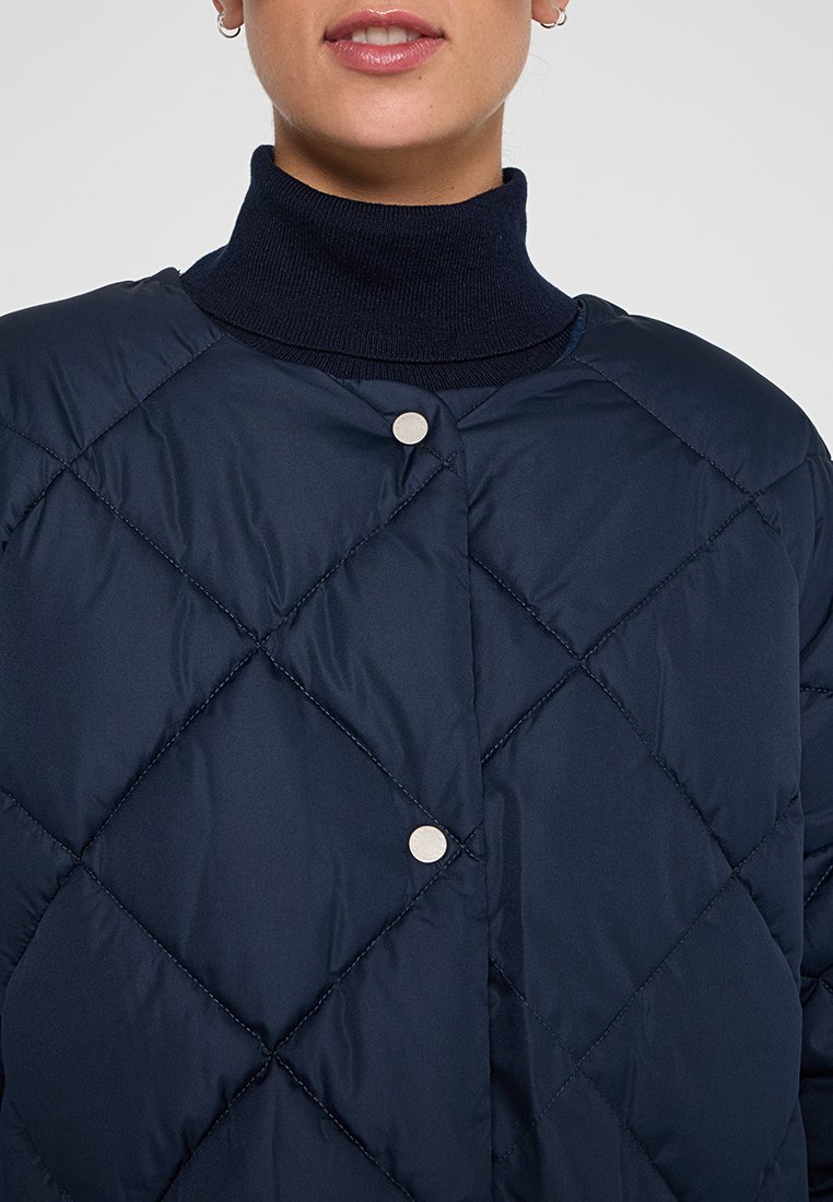 Granatowa pikowana kurtka z wysokim kołnierzem i metalowymi zatrzaskami. Tkanina ma gładką fakturę z wzorem w romby.