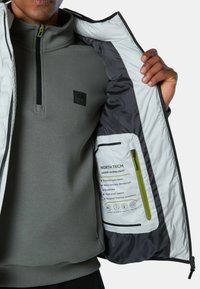 Grauer Pullover mit hohem Kragen, unter einer weißen Jacke. Die Jacke hat eine dunkle Innenauskleidung, einen grünen Reißverschluss und ein Innenetikett mit Pflegehinweisen.