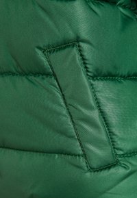 Zelená péřová bunda vyrobená z látky s hladkým, lesklým povrchem. Obsahuje obdélníkovou nášivku a prošívané švy pro zateplení.