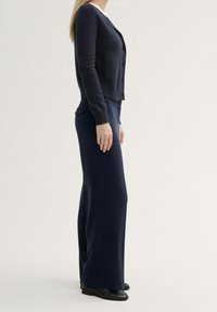 Marinefarbener Strickcardigan mit Knopfverschluss, kombiniert mit marinefarbener Anzughose mit Seitentaschen und geradem Schnitt. Schwarze Schuhe vervollständigen das Outfit.