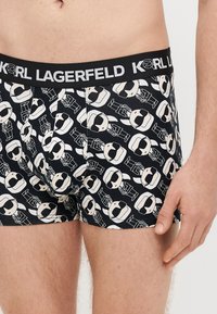 Melnas bokseršorti ar atkārtotu zīmējumu, kur attēlotas karikatūru figūras ar saulesbrillēm. Elastīgs jostasvietas lentes ar uzrakstu "KARL LAGERFELD" baltā krāsā.