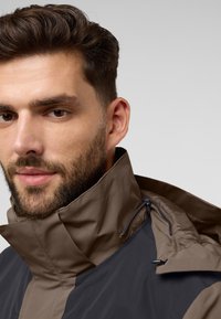 Waterdichte, tweekleurige jas met een bruine hoge kraag, ritssluitingen en een strakke, moderne pasvorm. Voorzijde met rits en textuurdetails in de stof.