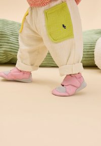 Rosa und graue weiche Schuhe mit Klettverschluss, kombiniert mit cremefarbenen gemusterten Hosen, die eine grüne Akzenttasche haben. Hintergrund in sanften Pastellfarben.