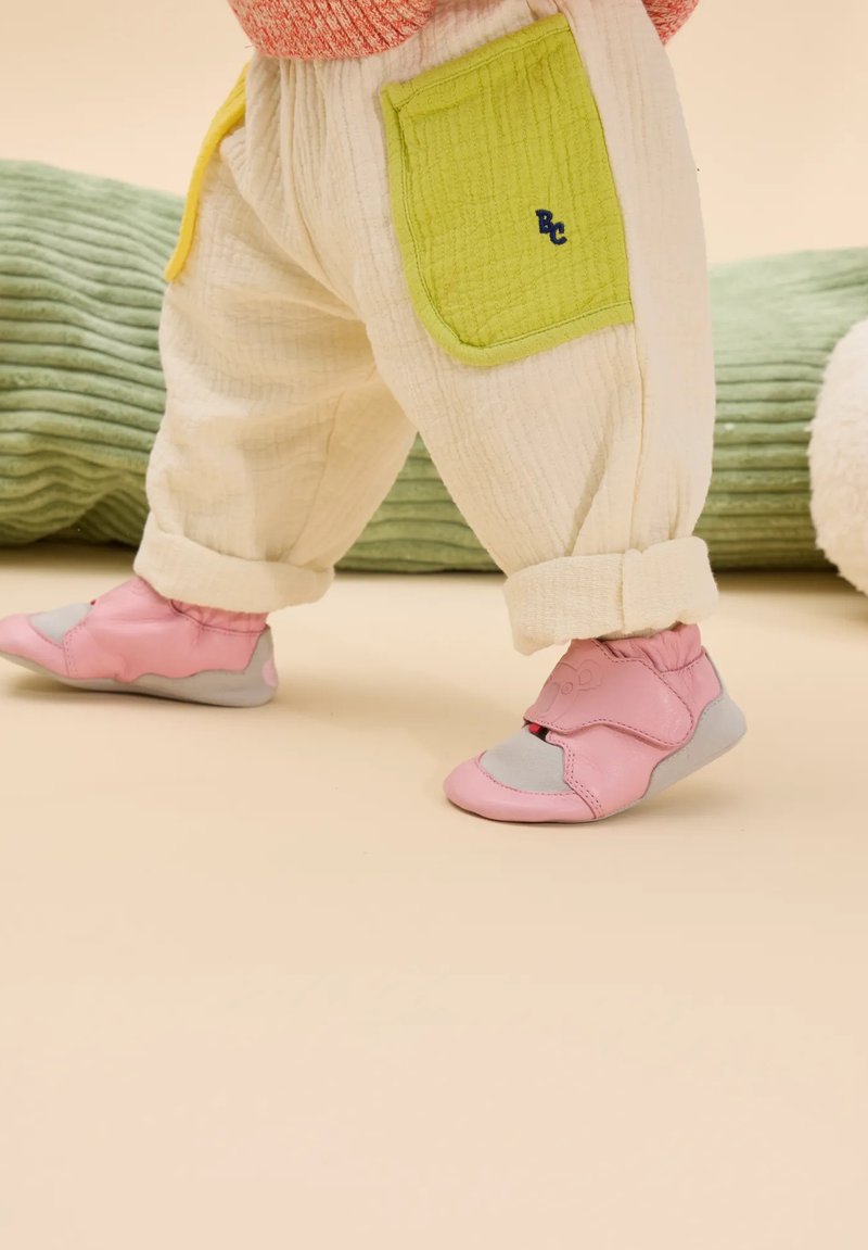 Rosa und graue weiche Schuhe mit Klettverschluss, kombiniert mit cremefarbenen gemusterten Hosen, die eine grüne Akzenttasche haben. Hintergrund in sanften Pastellfarben.