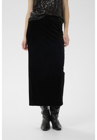 Juoda aksominė maksi sijonas su lygiu tekstūra, aptempiantis dizainas ir šoninės skeltukai. Dėvimas su juodais, iki kelių siekiančiais batais.