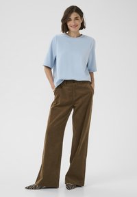 Žena stojaca s rukami v vreckách, oblečená v svetlomodrom krátkym rukávom, hnedých širokých nohaviciach a topánkach so zebrovým vzorom.