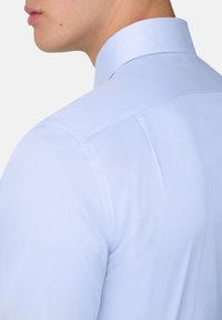 Nahaufnahme vom Hals und der Schulter einer Person, die ein hellblaues, fein gestreiftes Hemd mit steifem Kragen vor einem einfarbigen Hintergrund trägt.