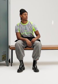 Szürke és neon zöld sportos póló "ETIHAD AIRWAYS" logóval, szürke bő nadrág, fekete bokacsizma, egy fa padon ülve.