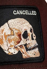 Aufnäher mit einem detaillierten, gestickten Schädelmotiv in Beige- und Brauntönen auf schwarzem Hintergrund, darüber das Wort "CANCELLED".