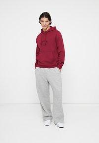 Burgundy hooded sweatshirt with a front pocket and black logo, paired with light grey sweatpants featuring side stripes and elastic cuffs.