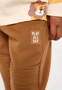 Braune Baumwoll-Jogginghosen mit strukturierten Kniefeldern und einem gewebten Etikett mit der Aufschrift "SPIEL DEN GANZEN TAG". Das Oberteil zeigt ein verspieltes Löwendesign.