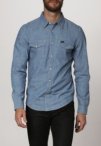 Vaaleansininen farkkupaita, jossa pitkät hihat, napillinen kaulus, kaksi rintataskua ja pieni brändipatch vasemmassa taskussa.