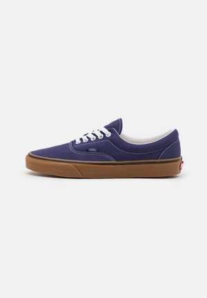 Niskie fioletowe trampki Vans z białymi sznurówkami i przeszyciami, wyposażone w brązową gumową podeszwę i logo Vans po boku i na pięcie.