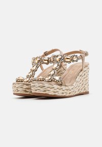Sandales compensées beiges avec une base en paille tissée, ornées de pierres précieuses claires et dorées sur la bride en T. Boucle réglable à la cheville incluse.