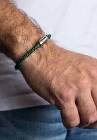 Žalias pintas apyrankė su nerūdijančio plieno sagtimi, uždėta ant riešo su šviesiais plaukais. Fone matoma šviesi marškiniai ir džinsai.