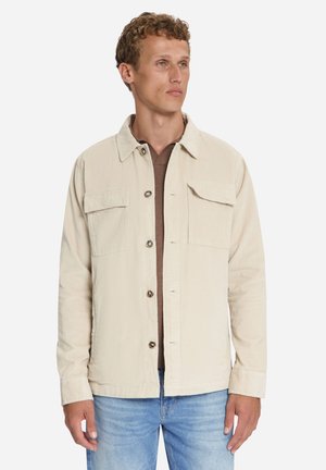 Junger Mann trägt eine beige, geknöpfte Jacke über einem braunen Hemd und hellblauen Jeans, schaut leicht nach rechts vor einem einfarbigen Hintergrund.