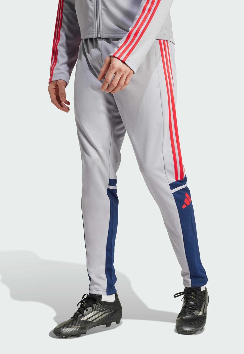Grey athletic pants with red and blue side stripes, made of smooth fabric. Features an elastic waistband and tapered leg design.