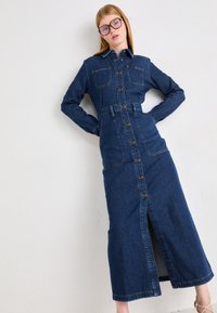 Vestido de mezclilla con mangas largas, botonera en la parte delantera, dos bolsillos en el pecho, cintura ajustada con cinturón, longitud hasta la mitad de la pantorrilla y una abertura central en la parte delantera.