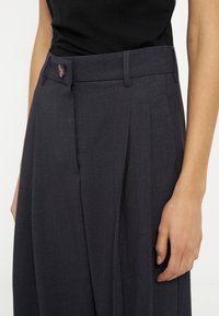 Gros plan sur une personne portant un pantalon ajusté gris foncé avec des plis et une fermeture à bouton, associé à un haut noir.