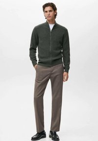 Cardigan verde a coste con zip frontale, abbinato a pantaloni marrone chiaro e scarpe nere. Tessuto morbido con design aderente e maniche lunghe.