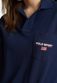 Gros plan d'une personne portant un polo bleu marine avec un col et une poche affichant "POLO SPORT Ralph Lauren" et un drapeau américain.