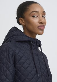 Mornarski jakna s kapuljačom, prošivena, s patentnim zatvaračem i vezicom. Tkanina ima glatku teksturu i dijamantni uzorak.