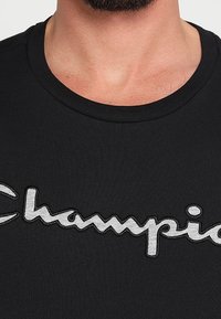 Schwarzes Baumwoll-T-Shirt mit Rundhalsausschnitt und einem auffälligen, grauen, gestickten "Champion"-Logo auf der Brust.