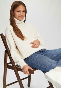 Cremfarbener gerippter Rollkragenpullover, lockere Passform, kombiniert mit blauen Jeans und weißen Sneakers. Modell sitzt auf einem Holzstuhl.