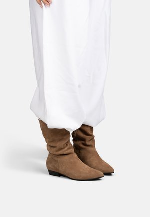Botas marrones de ante hasta la rodilla con tacón bajo, usadas con una prenda larga blanca, mostradas desde la mitad de la pantorrilla hacia abajo sobre un fondo claro.